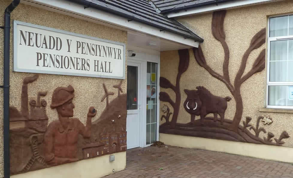 Miner holding coal in front of a scene of Ammanford with brickworks, rail line and wind turbines. And the Boar in a forest. All in cob with some detail in mosaic.